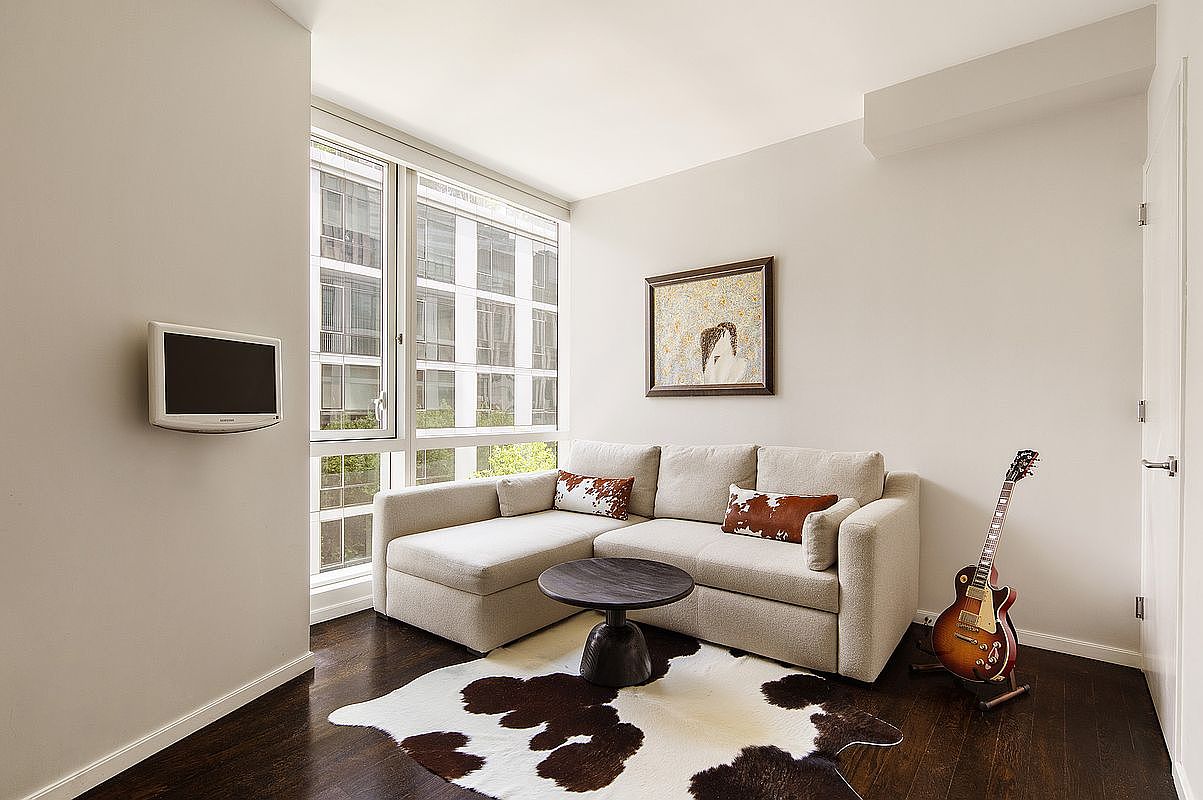 This is an interior shot of a living room featuring a beige sectional sofa with decorative pillows, a round coffee table, and a cowhide rug. A flat-screen TV is mounted on the wall, and a framed artwork hangs above the sofa. A guitar stands near the wall, adding a touch of personality to the space. Large windows provide natural light, and the dark wood flooring adds warmth.