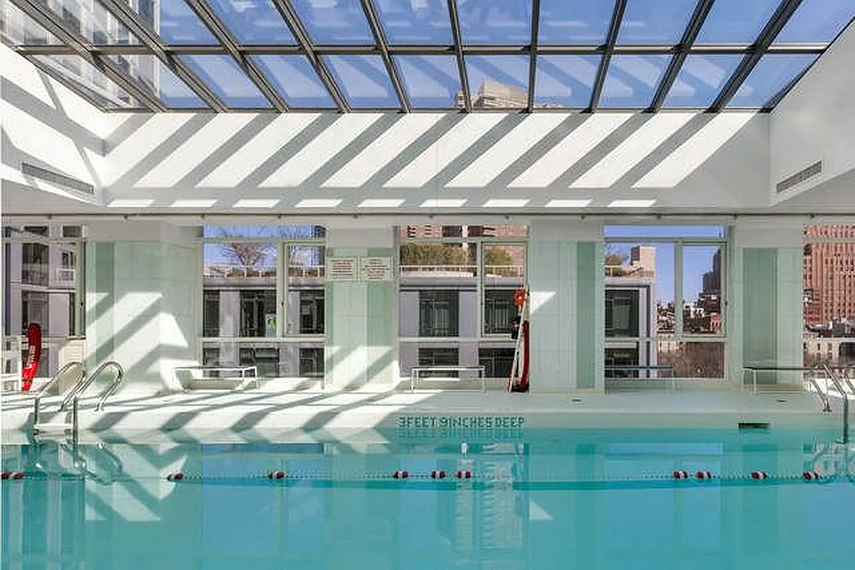 This image showcases a bright and inviting indoor swimming pool area, likely within a residential building or community amenity space. The pool features clear blue water, lane markers, and ample natural light streaming through a large glass ceiling. The surrounding area is clean and modern, suggesting a luxurious and well-maintained environment.