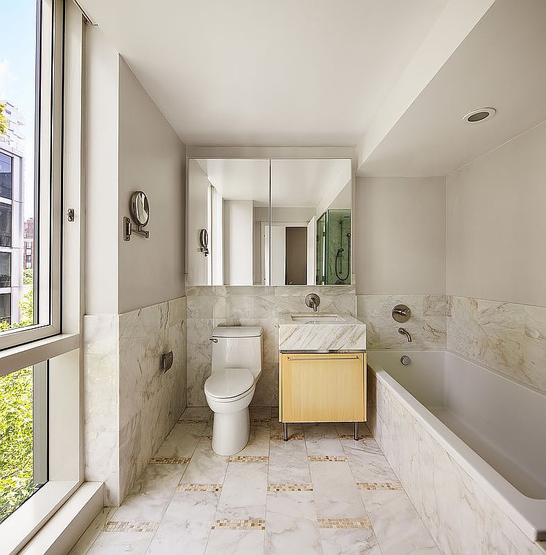 This is a well-lit primary bathroom featuring marble tiling on the walls and floor, creating a luxurious and clean aesthetic. The bathroom includes a toilet, a vanity with a marble countertop and light wood cabinetry, a large mirror, and a bathtub. A large window provides natural light, enhancing the bright and airy feel of the space.