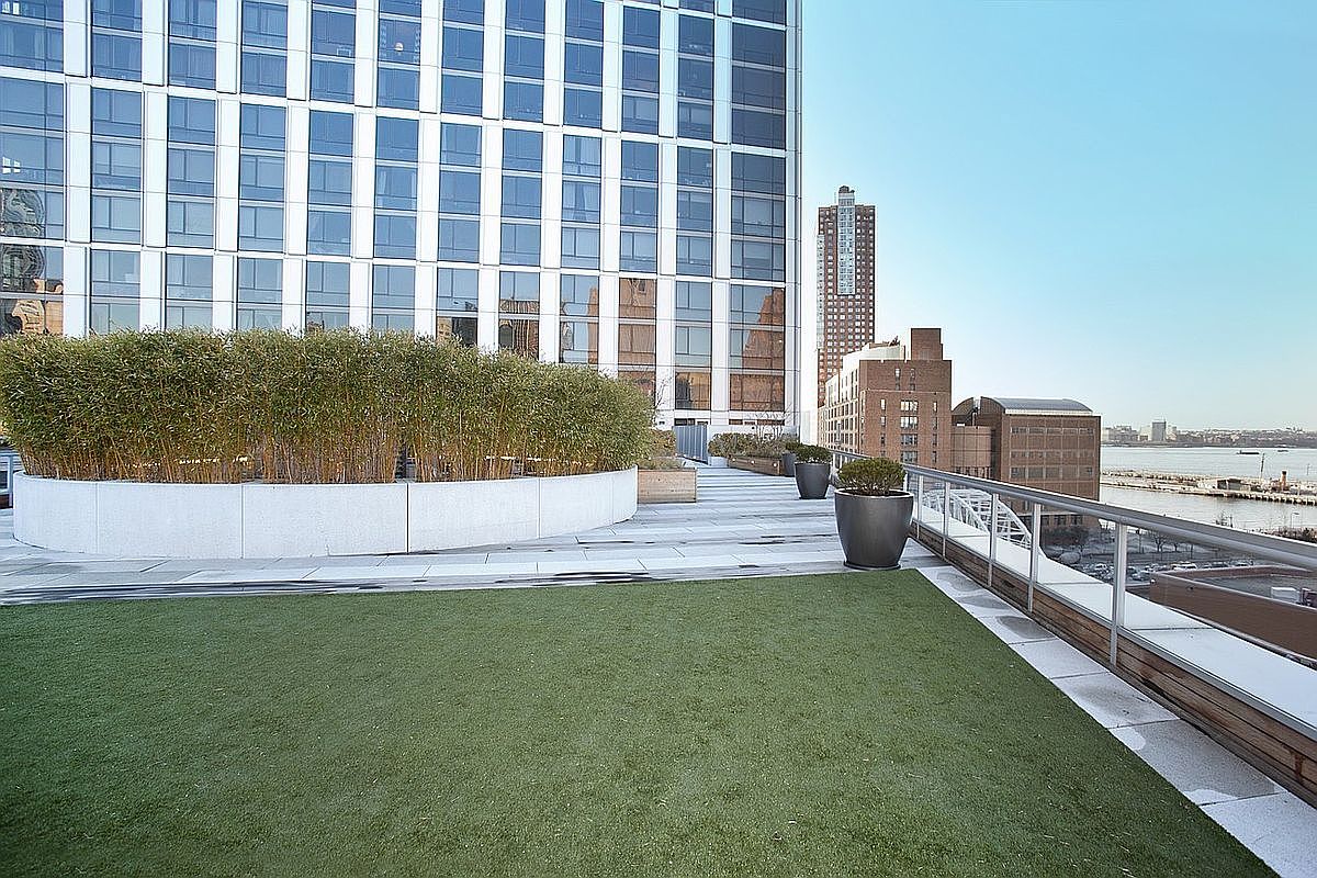 This image showcases a modern rooftop patio or balcony with artificial turf, planters with bamboo, and sleek railings. The backdrop features a contemporary building with large windows and other buildings in the distance, suggesting an urban setting. The space appears well-maintained and offers a private outdoor area with city views.