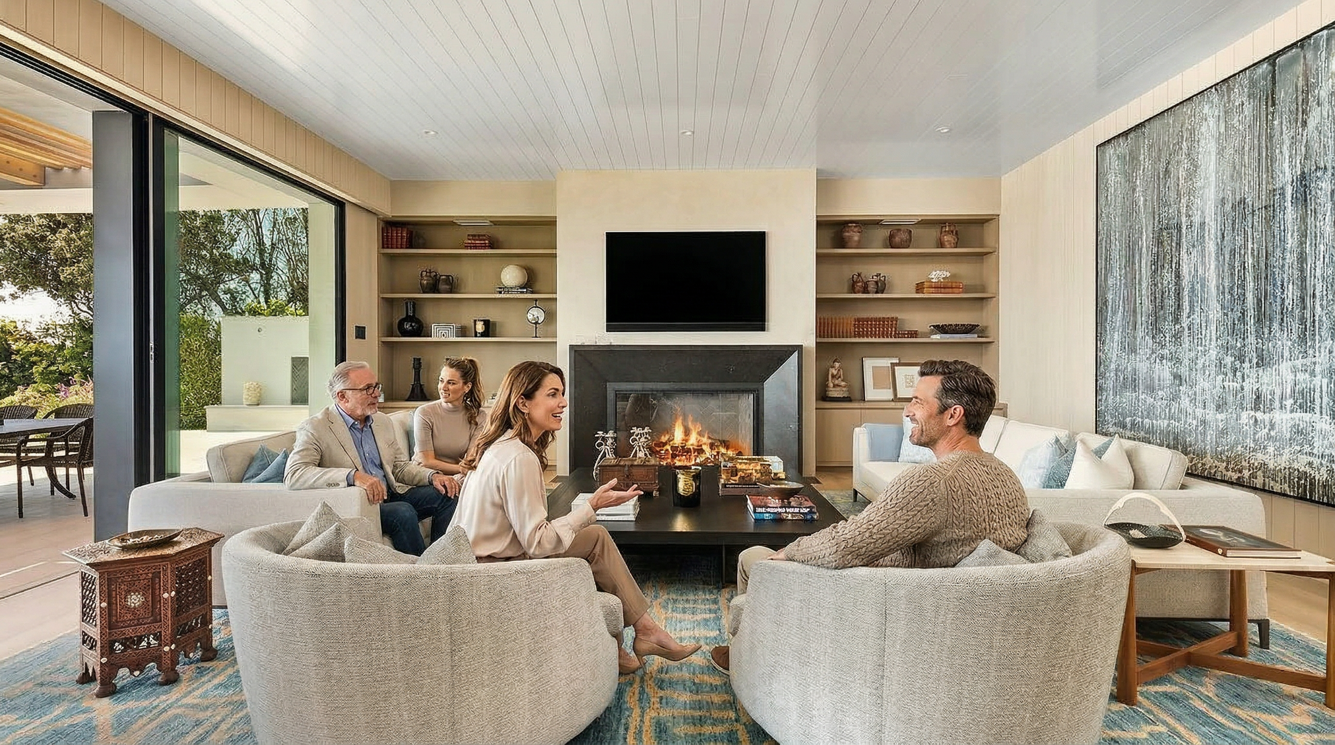 This sophisticated living room features a symmetrical layout with two plush white sofas facing each other across a large, dark wood coffee table. A central fireplace with a mounted television serves as the focal point, flanked by built-in shelving units filled with curated decor. The space is anchored by a vibrant blue and gold patterned rug, while floor-to-ceiling glass doors provide a seamless transition to the outdoor patio, creating an airy and luxurious atmosphere.
