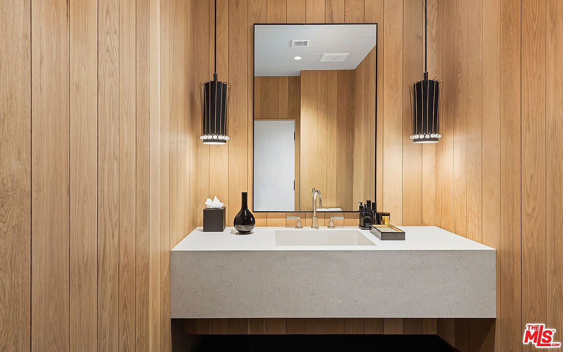 This elegant powder room features warm, vertical wood-paneled walls that create a sophisticated and organic atmosphere. A minimalist white stone vanity with an integrated sink is centered beneath a sleek, black-framed mirror, flanked by two modern, cage-style pendant lights. The space is clean and contemporary, offering a high-end, spa-like aesthetic perfect for a guest bathroom.
