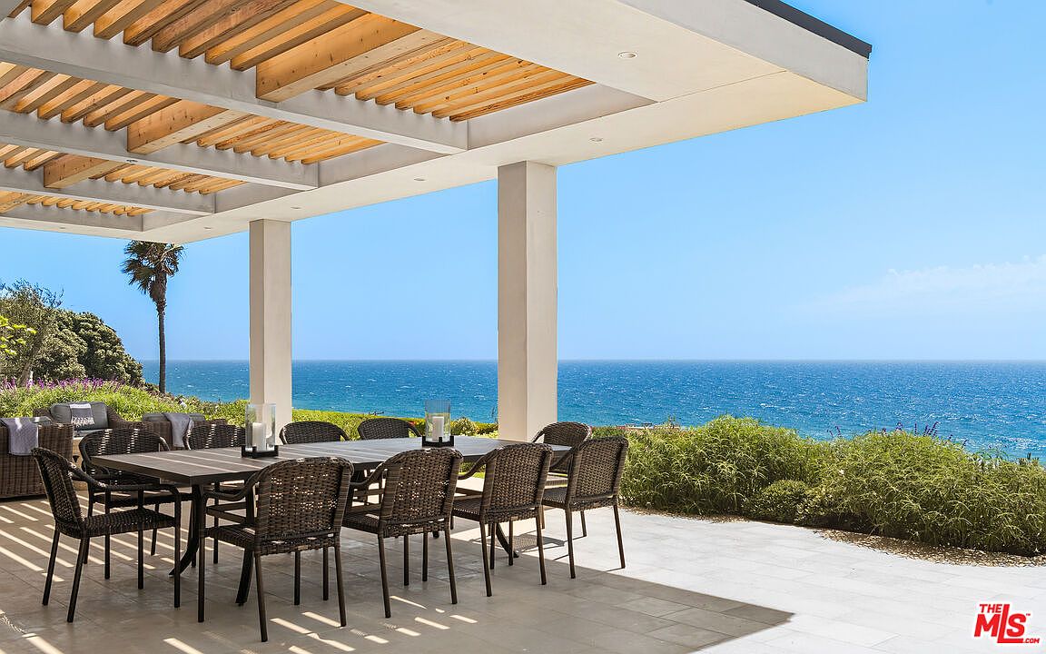 This stunning outdoor patio features a large dining set under a modern pergola with wooden slats, offering a seamless transition to an expansive ocean view. The space is paved with light-colored stone tiles and surrounded by lush greenery, creating a serene and luxurious atmosphere perfect for al fresco dining. The perspective captures the open-air design, emphasizing the property's prime coastal location and sophisticated architectural style.