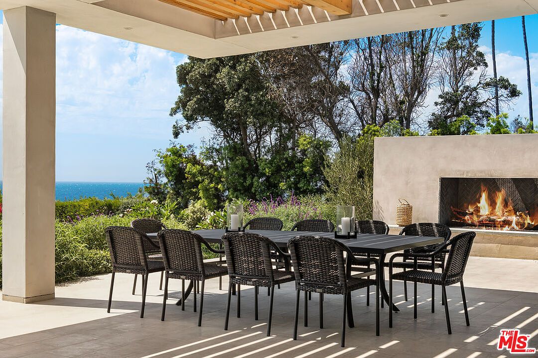 This elegant outdoor patio features a large dining table with wicker chairs, perfectly positioned for al fresco dining with a stunning ocean view. The space is anchored by a modern, built-in fireplace set into a clean-lined concrete wall, creating a sophisticated and cozy atmosphere. The open-air design, combined with the lush greenery and coastal backdrop, offers a luxurious and serene setting for entertaining.