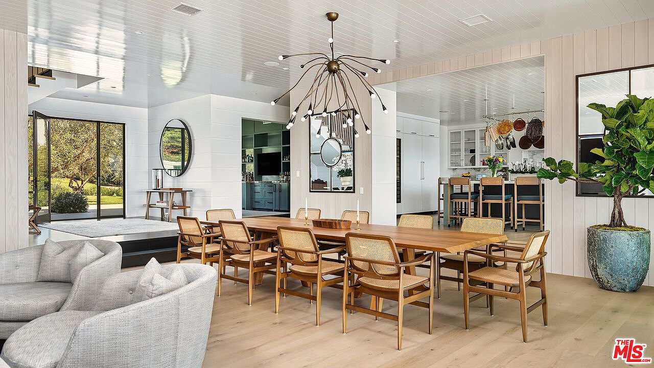 This elegant dining room features a long, natural wood table surrounded by mid-century modern cane-back chairs, positioned beneath a striking, multi-armed sputnik-style chandelier. The open-concept space flows seamlessly into a bright kitchen and a cozy living area, all tied together by light-toned wood flooring and crisp white shiplap walls. A large potted fiddle leaf fig adds a touch of organic warmth to the sophisticated, airy interior.
