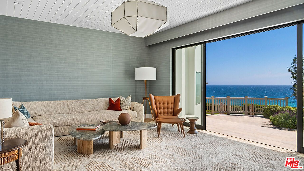 This sophisticated living room features a neutral-toned sofa, a unique marble-topped coffee table, and a stylish tan leather armchair, all set against a textured blue-grey accent wall. Large sliding glass doors open directly onto a spacious deck, providing a seamless transition to a breathtaking, unobstructed view of the ocean. The room is illuminated by a modern geometric pendant light and a floor lamp, creating an airy, luxurious atmosphere perfect for coastal living.