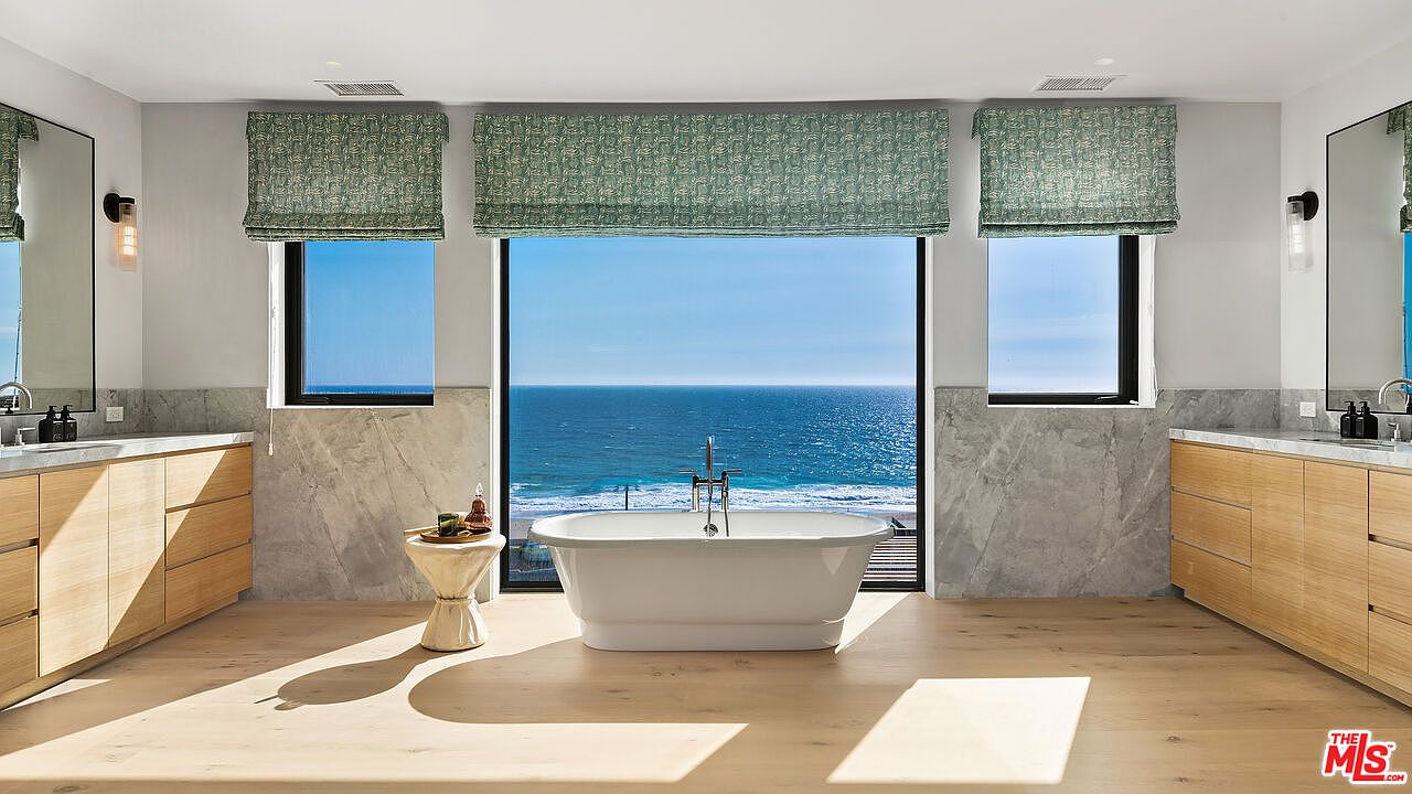 This luxurious primary bathroom features a minimalist, modern aesthetic with a central freestanding soaking tub positioned perfectly in front of a large floor-to-ceiling window overlooking the ocean. The space is anchored by dual light-wood vanities with marble countertops and matching wall-mounted mirrors, creating a symmetrical and serene atmosphere. The combination of natural wood tones, cool marble, and the expansive coastal view provides a high-end, spa-like experience.