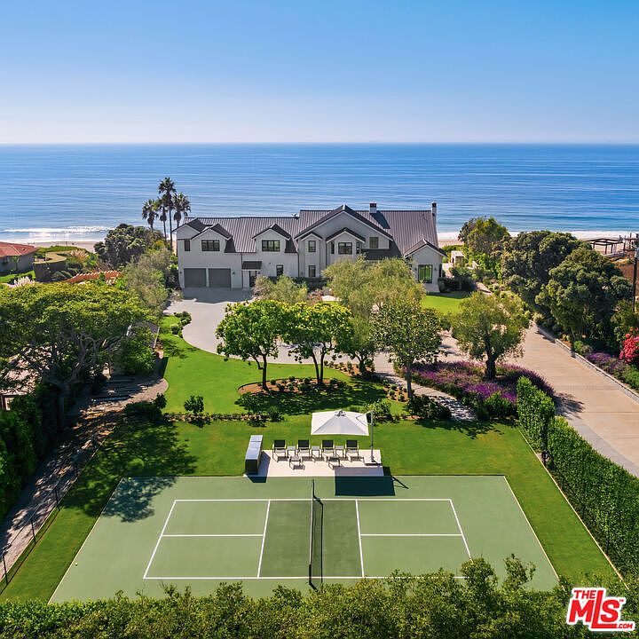 This high-angle aerial shot captures a sprawling coastal estate featuring a large, multi-gabled residence situated directly overlooking the ocean. The property boasts an expansive, meticulously landscaped lawn that includes a private tennis court and a dedicated outdoor seating area with a large umbrella. The scene conveys a sense of luxury and tranquility, highlighting the seamless integration of high-end residential living with a stunning beachfront environment.