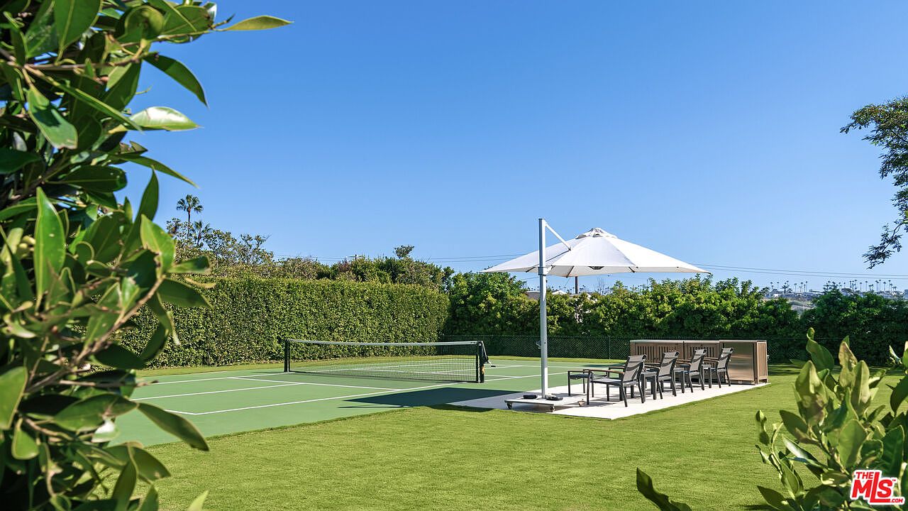 This outdoor space features a well-maintained tennis court bordered by lush green hedges and a manicured lawn. A large, modern cantilevered umbrella shades a patio dining set, creating a perfect area for relaxation or social gatherings. The bright, sunny atmosphere and clear blue sky emphasize the property's expansive and private recreational amenities.