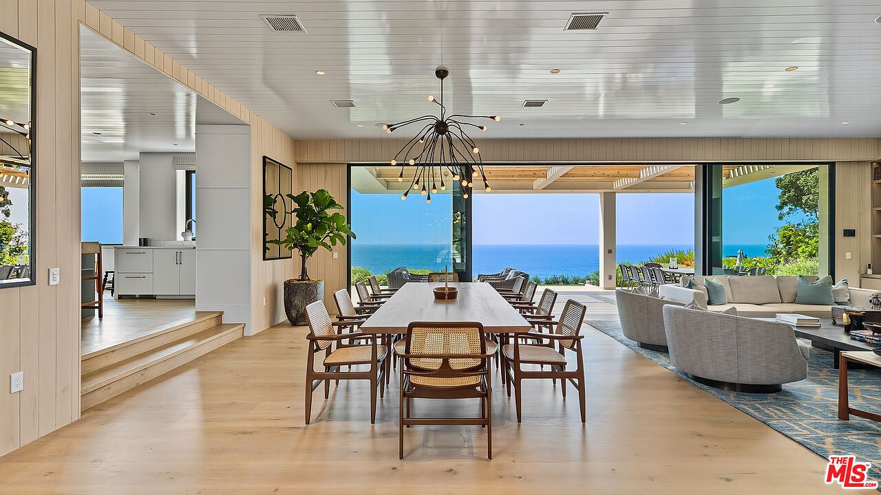 This elegant open-concept dining area features a long wooden table surrounded by mid-century modern cane-back chairs, illuminated by a striking sputnik-style chandelier. The space seamlessly transitions into a living area and opens up to a breathtaking panoramic view of the ocean through expansive floor-to-ceiling glass walls. The interior design emphasizes a light, airy aesthetic with natural wood tones and a sophisticated, coastal-modern atmosphere.