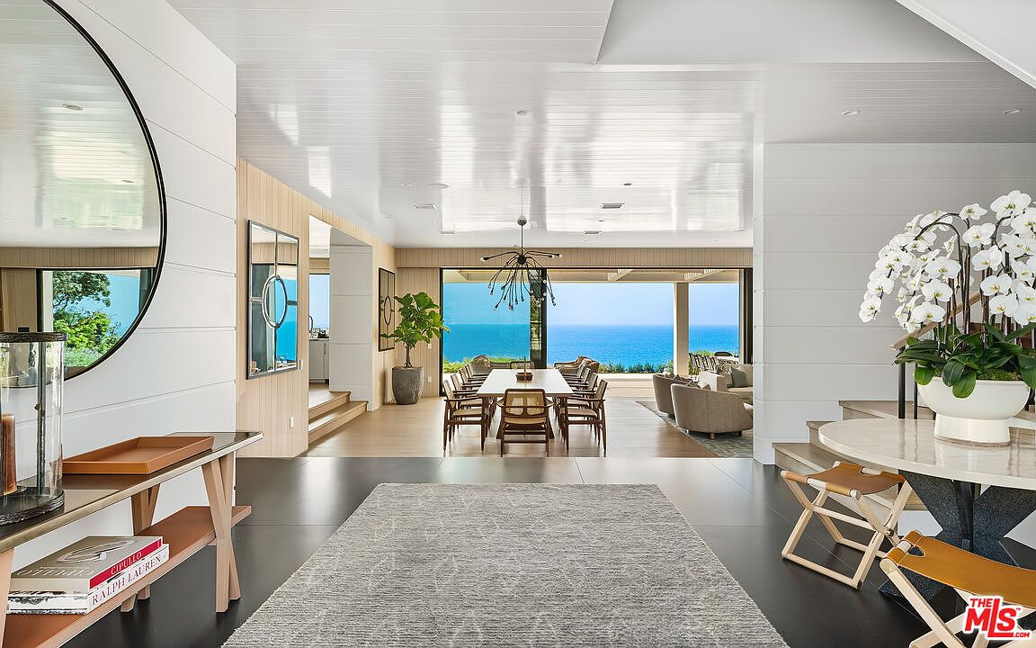 This elegant entryway transitions into a spacious, open-concept living and dining area that frames a breathtaking panoramic view of the ocean. The interior features a sophisticated, coastal-modern aesthetic with crisp white shiplap walls, dark flooring, and high-end furnishings, including a round marble table and a sleek dining set. The perspective is centered, drawing the eye through the home toward the expansive glass doors that seamlessly connect the indoor living space to the outdoor horizon.