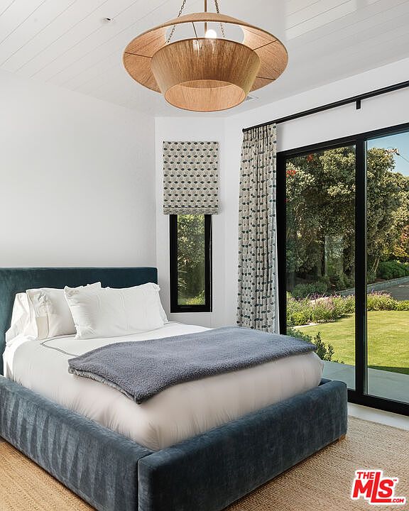 This serene bedroom features a plush, dark blue upholstered bed frame topped with a cozy grey throw blanket and crisp white linens. A unique, tiered wooden pendant light serves as a focal point, while floor-to-ceiling glass sliding doors provide a seamless connection to the lush green backyard. The room is accented by a patterned Roman shade on a narrow window, creating a clean, modern, and inviting atmosphere.