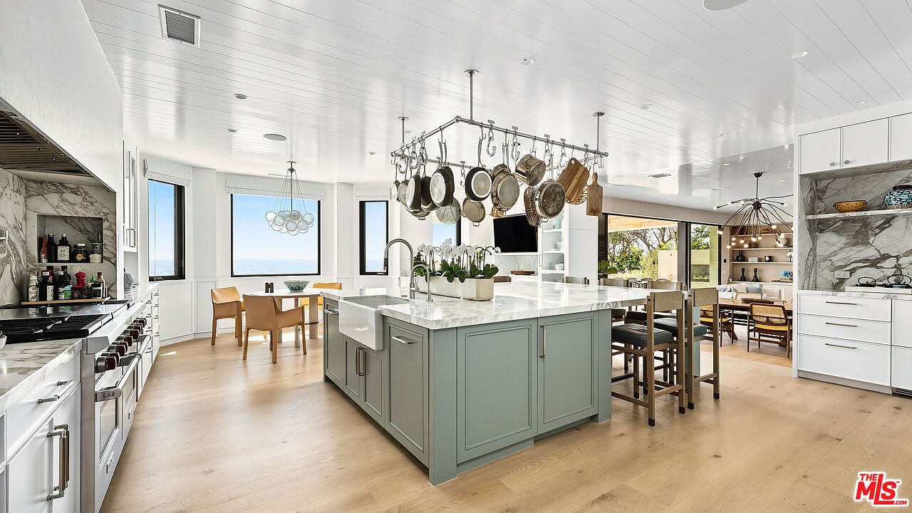 This bright and airy kitchen features a large, sage-green central island with a marble countertop and a farmhouse sink, serving as the focal point of the room. A prominent overhead pot rack hangs above the island, while white cabinetry and light wood flooring create a clean, coastal-inspired aesthetic. The space seamlessly transitions into a dining area with ocean views, emphasizing an open-concept layout perfect for modern living.