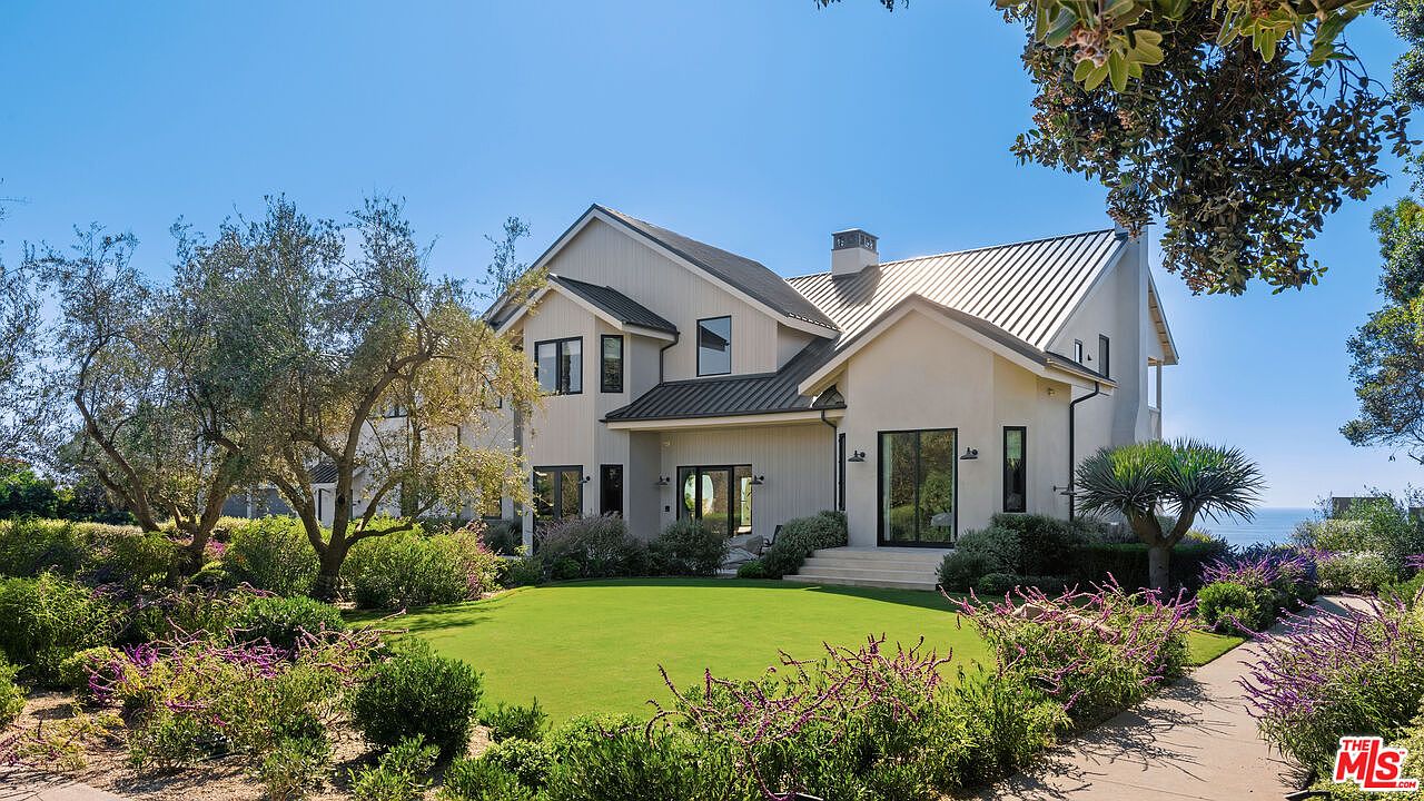 This stunning rear view of a modern farmhouse-style residence showcases a clean, light-colored exterior with a striking dark metal roof and large, black-framed windows. The home opens onto a meticulously manicured green lawn, framed by vibrant purple flowering shrubs and mature trees, creating a serene and inviting outdoor living space. The perspective captures the architectural elegance of the home against a clear blue sky, with a glimpse of the ocean in the distance, suggesting a luxurious coastal lifestyle.