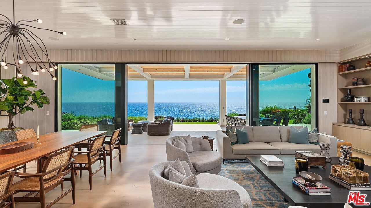 This expansive, open-concept living and dining area features a seamless transition to an outdoor patio through floor-to-ceiling glass sliding doors that frame a stunning ocean view. The interior is decorated in a sophisticated, neutral palette with contemporary furnishings, including a plush sofa, curved armchairs, and a long wooden dining table. The space is illuminated by a striking modern chandelier, creating an airy, luxurious atmosphere perfect for coastal living.