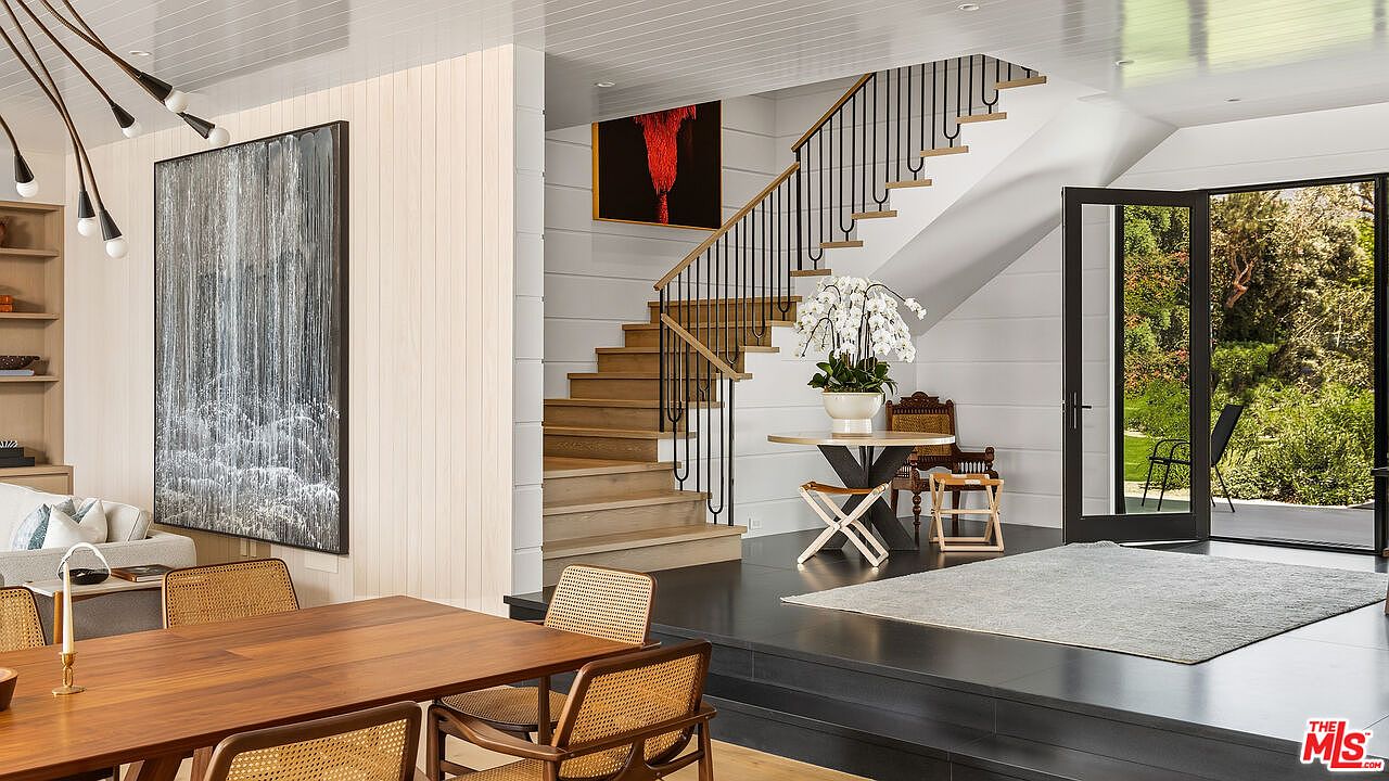 This elegant entryway features a striking staircase with light wood treads and a sleek black metal railing, serving as the focal point of the space. To the left, a large abstract art piece hangs above a dining area, while a raised platform leads to a bright, open doorway that connects the interior to the outdoor patio. The design blends modern architectural elements with warm, natural textures, creating a sophisticated and inviting atmosphere.