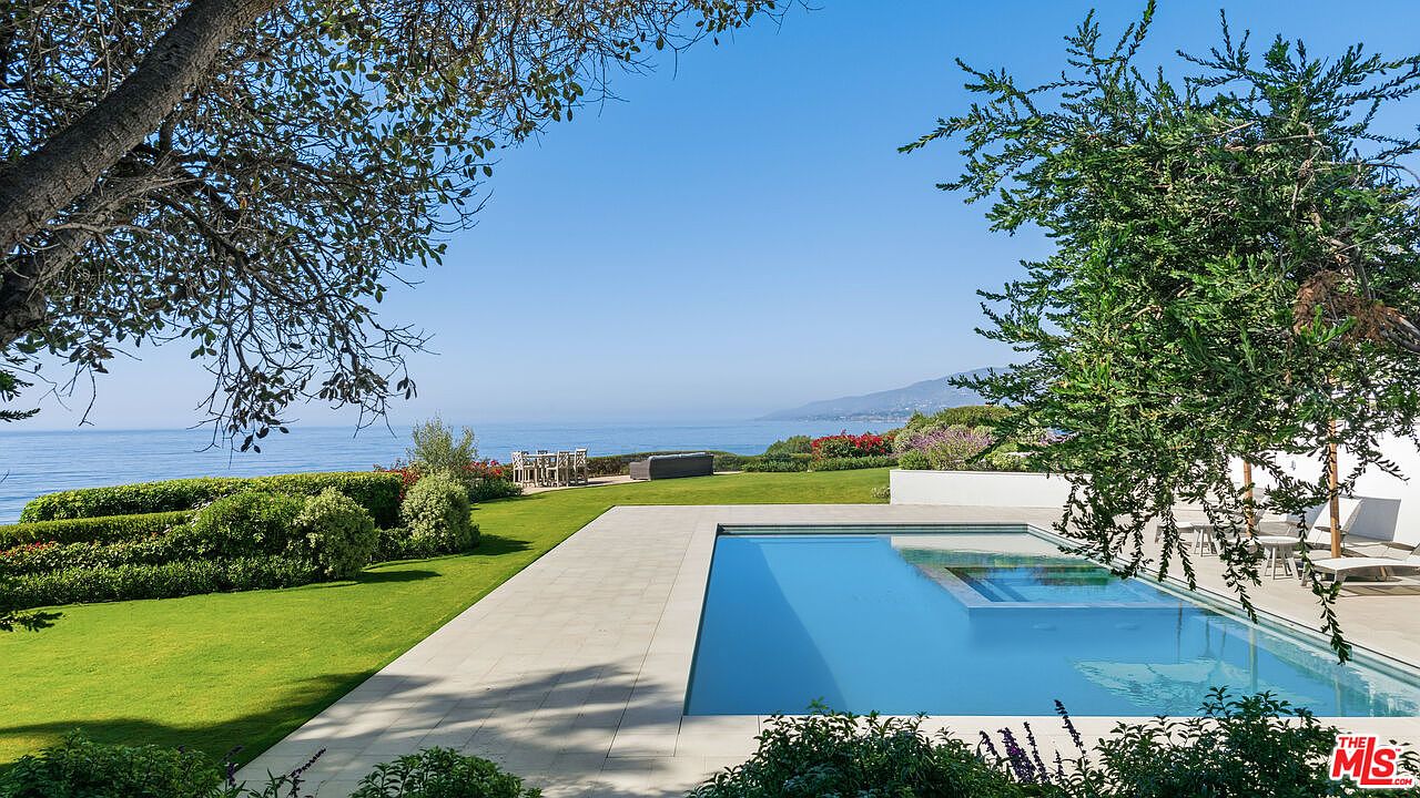 This stunning outdoor space features a sleek, rectangular infinity-edge pool with an integrated spa, perfectly positioned to overlook the vast ocean horizon. The manicured lawn and lush landscaping frame the scene, while a patio area with outdoor seating provides a serene spot for relaxation. The perspective captures the seamless blend of luxury architecture with the natural beauty of the coastal landscape.