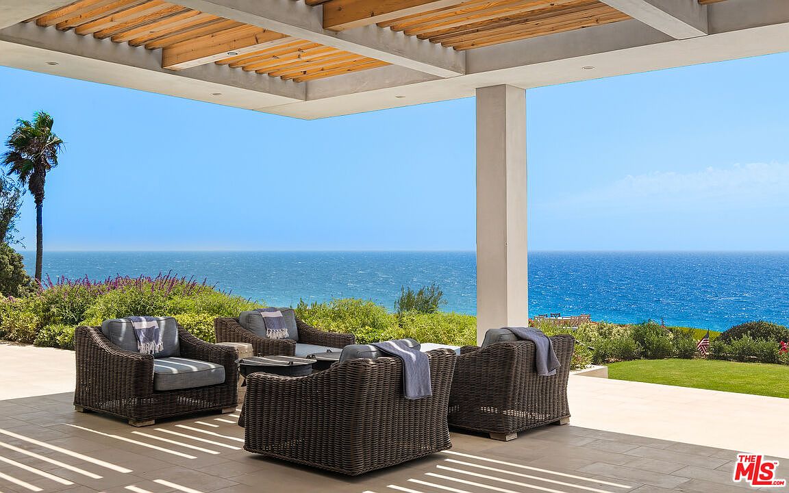 This expansive covered patio offers a luxurious outdoor living space with comfortable wicker lounge seating arranged to face the ocean. The area features a modern pergola ceiling and sleek stone flooring, seamlessly blending indoor comfort with breathtaking, unobstructed panoramic views of the sea. The setting is serene and sophisticated, perfect for high-end coastal relaxation.