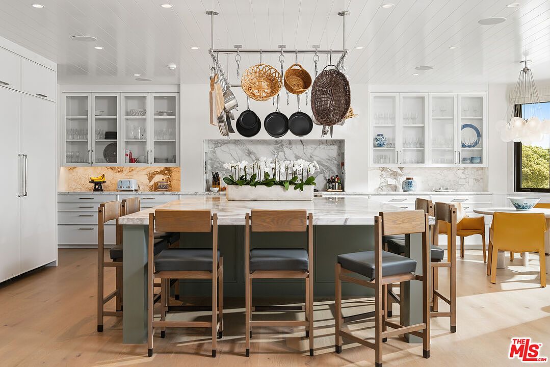 This bright and airy kitchen features a large central island with a marble countertop and four wooden bar stools, serving as the focal point of the room. Above the island, a hanging pot rack displays various cookware and woven baskets, while white cabinetry and marble backsplashes provide a clean, sophisticated aesthetic. The space is illuminated by recessed lighting and a modern chandelier, creating a welcoming and luxurious atmosphere perfect for culinary activities.