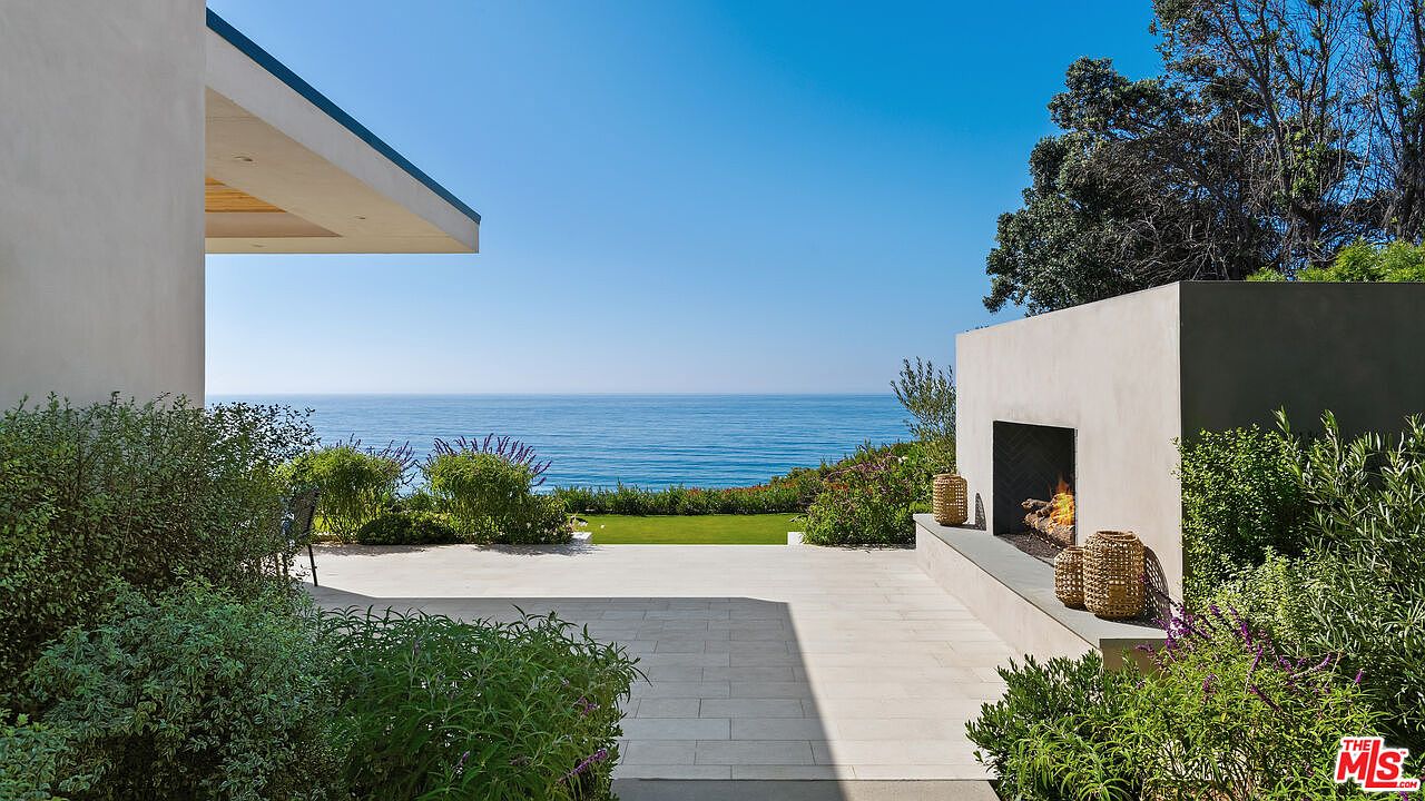 This stunning outdoor patio space features a sleek, modern fireplace built into a minimalist concrete wall, overlooking a breathtaking panoramic ocean view. The area is paved with light-colored stone tiles and framed by lush, manicured greenery, creating a serene and luxurious atmosphere. The perspective captures the seamless transition between the architectural structure and the expansive horizon, emphasizing the property's prime coastal location.
