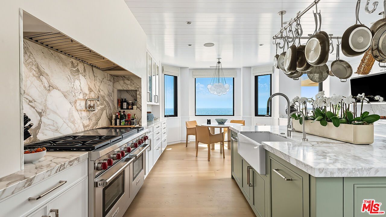 This bright and airy kitchen features a high-end Wolf range, marble countertops, and a large farmhouse sink set into a sage green island. A hanging pot rack adds a professional culinary touch, while the open-concept layout flows seamlessly toward a dining nook with panoramic ocean views. The space is defined by its clean, coastal-inspired aesthetic, combining luxury finishes with a serene, sun-drenched atmosphere.