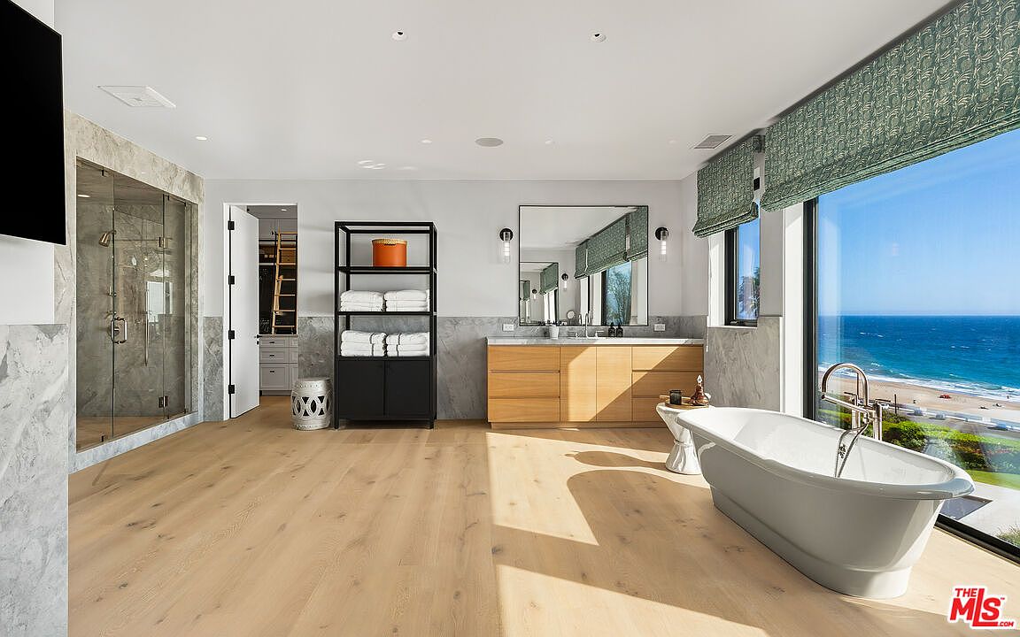 This luxurious primary bathroom features a sleek, modern design with light wood flooring and a freestanding soaking tub positioned to overlook a stunning ocean view. The space includes a double vanity with a large mirror, a glass-enclosed walk-in shower, and a minimalist shelving unit, all bathed in natural light. The overall aesthetic is serene, high-end, and perfectly integrated with the coastal surroundings.