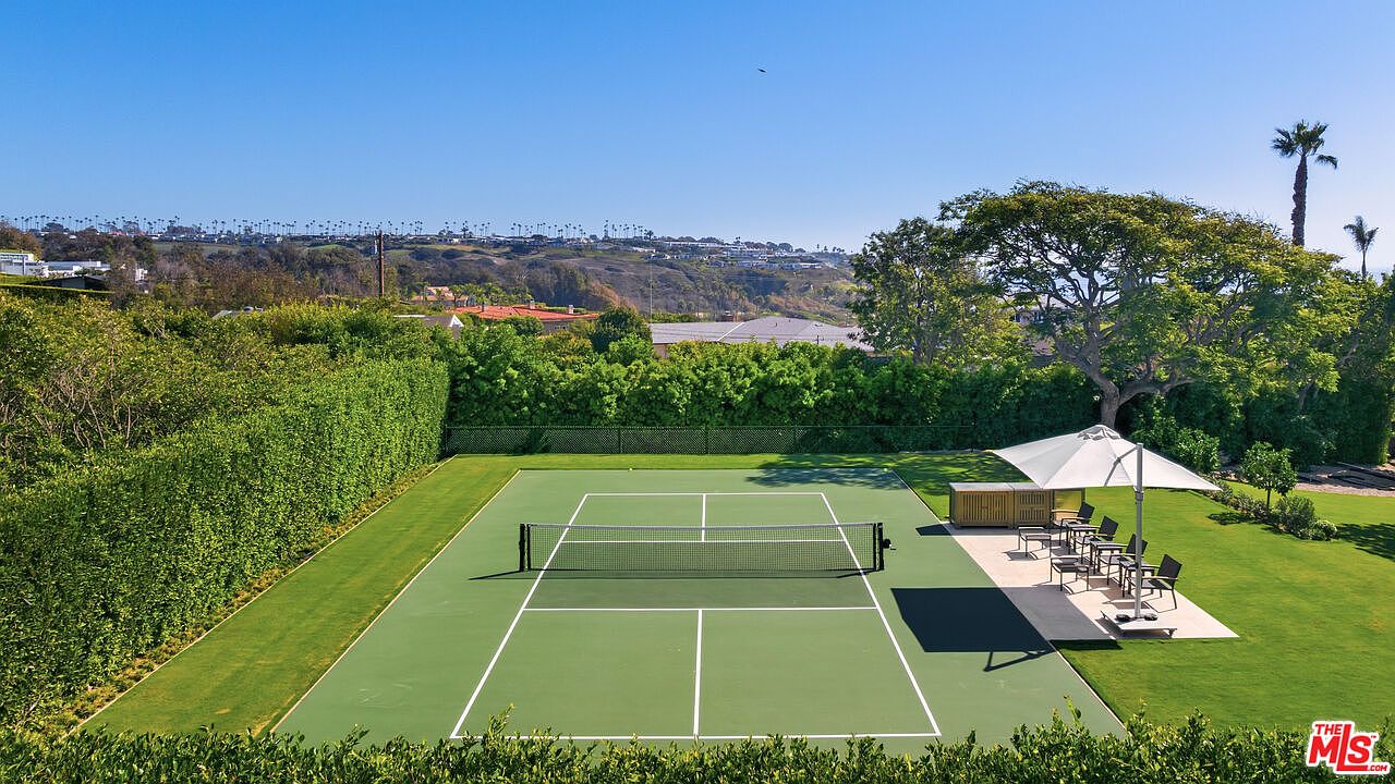 This elevated, high-angle shot showcases a pristine private tennis court nestled within a lush, manicured backyard. The scene features a vibrant green court surface, a shaded seating area with an umbrella, and a dense perimeter of tall, mature hedges that provide excellent privacy. In the background, rolling hills and a distant residential community create a scenic, expansive backdrop under a clear blue sky.