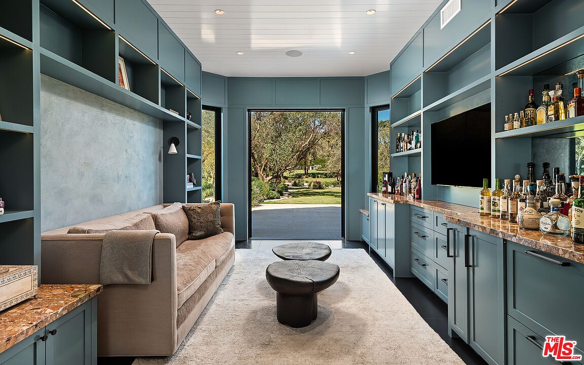 This sophisticated lounge area features custom built-in teal cabinetry with integrated lighting, a plush beige sofa, and a central pair of dark, organic-shaped coffee tables. The room is anchored by a soft, light-colored rug and opens directly to a lush, sunlit garden through a large glass doorway. The space exudes a refined, modern aesthetic, perfectly blending cozy relaxation with a high-end home bar setup.