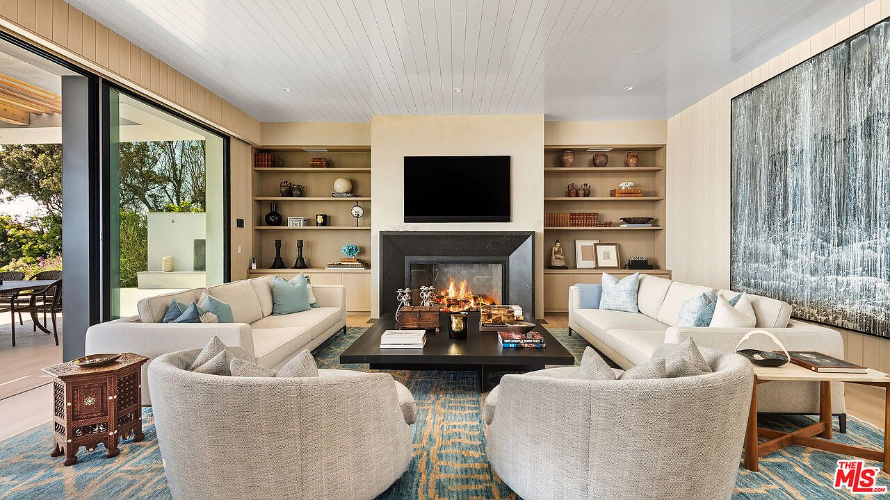 This sophisticated living room features a symmetrical layout with two plush white sofas facing each other across a large, dark wood coffee table. A central fireplace with a mounted television serves as the focal point, flanked by built-in shelving units filled with curated decor. The space is anchored by a vibrant blue and gold patterned rug, while floor-to-ceiling glass doors provide a seamless transition to the outdoor patio, creating an airy and luxurious atmosphere.