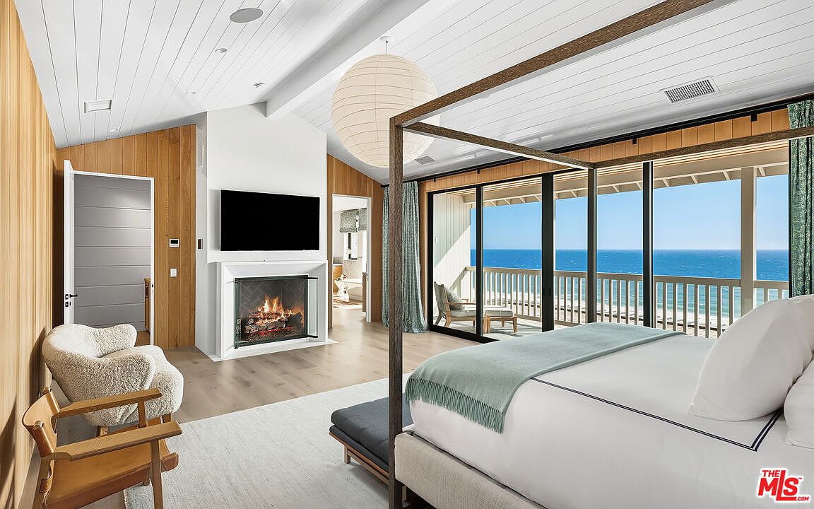 This luxurious primary bedroom features a sophisticated coastal aesthetic with light wood paneling, a vaulted ceiling, and a cozy fireplace integrated into a white accent wall. A four-poster bed serves as the centerpiece, positioned to take full advantage of the expansive floor-to-ceiling glass doors that reveal a stunning, unobstructed view of the ocean. The room is bathed in natural light, creating a serene and airy atmosphere that seamlessly blends indoor comfort with the beauty of the seaside landscape.
