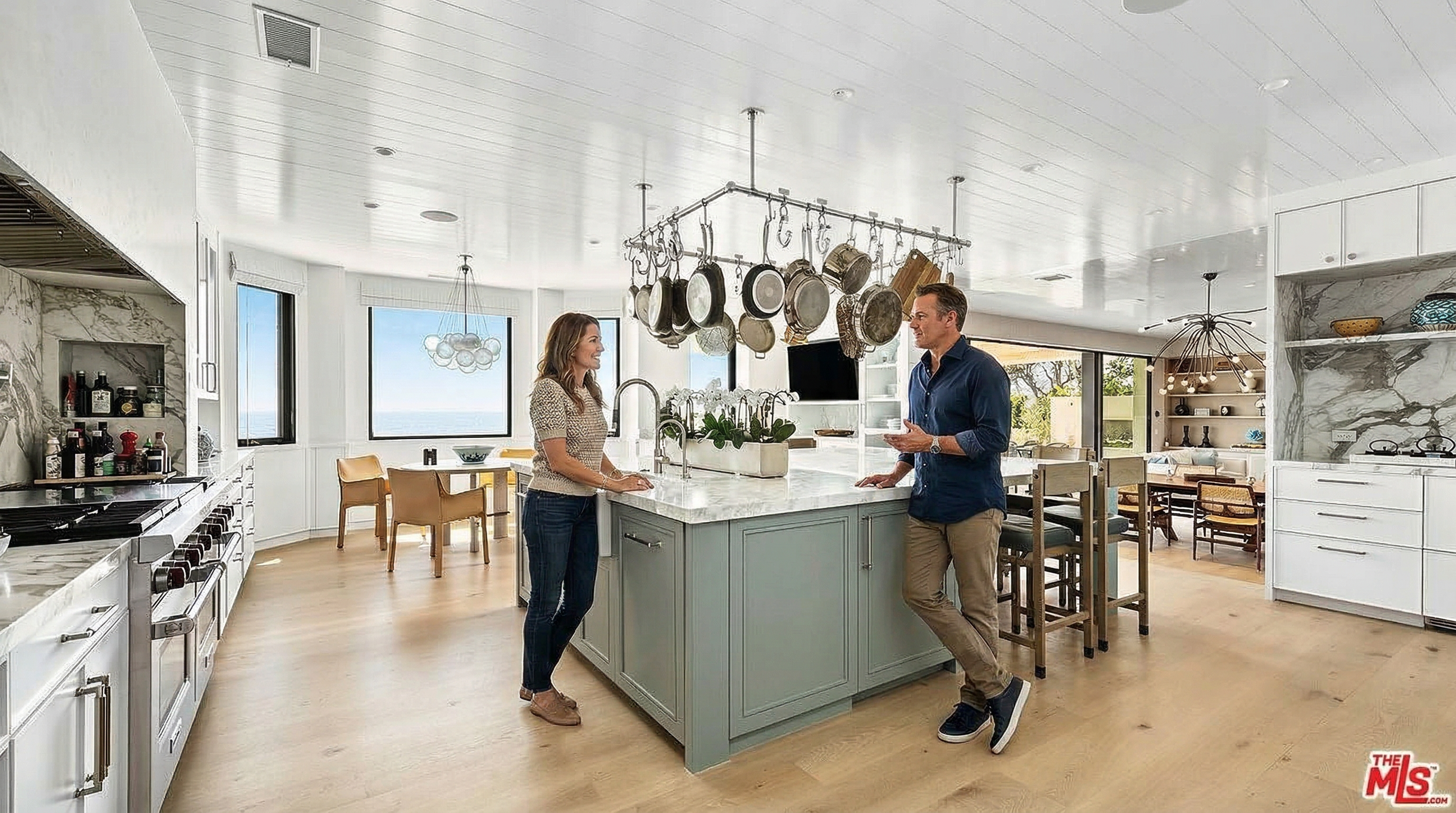 This bright and airy kitchen features a large, sage-green central island with a marble countertop and a farmhouse sink, serving as the focal point of the room. A prominent overhead pot rack hangs above the island, while white cabinetry and light wood flooring create a clean, coastal-inspired aesthetic. The space seamlessly transitions into a dining area with ocean views, emphasizing an open-concept layout perfect for modern living.