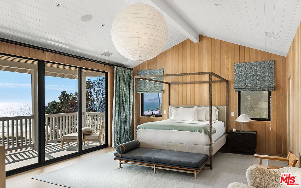 This serene primary bedroom features a sophisticated canopy bed as the centerpiece, set against warm, vertical wood-paneled walls. Large sliding glass doors open onto a private balcony with stunning ocean views, while a large paper lantern pendant light adds a soft, modern touch to the vaulted ceiling. The room is elegantly furnished with a dark upholstered bench and a minimalist nightstand, creating a tranquil and luxurious coastal retreat.