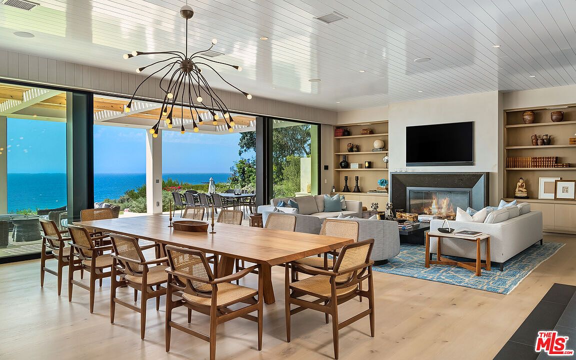 This expansive, open-concept living and dining area features a seamless transition to an outdoor patio with stunning ocean views through floor-to-ceiling glass walls. The space is anchored by a modern fireplace with a large television above, flanked by custom built-in shelving, and illuminated by a striking, sculptural sputnik-style chandelier. The interior design blends natural wood tones, light-colored upholstery, and a textured blue area rug to create a sophisticated, airy, and coastal-inspired atmosphere.