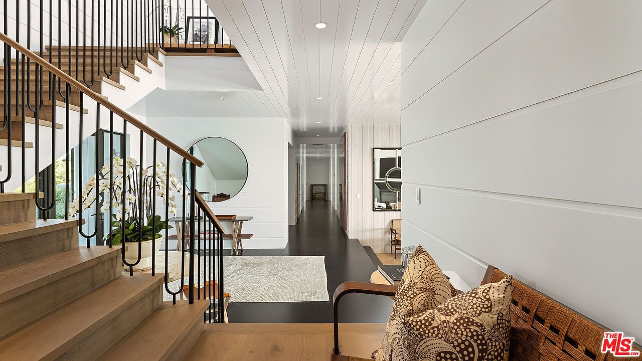This elegant entryway features a striking staircase with light wood treads and a sleek black metal railing, leading to an upper landing. The space is characterized by crisp white shiplap walls, a long hallway with dark flooring, and a large circular mirror that adds a modern touch. The bright, airy atmosphere is enhanced by recessed lighting and a clean, minimalist aesthetic.