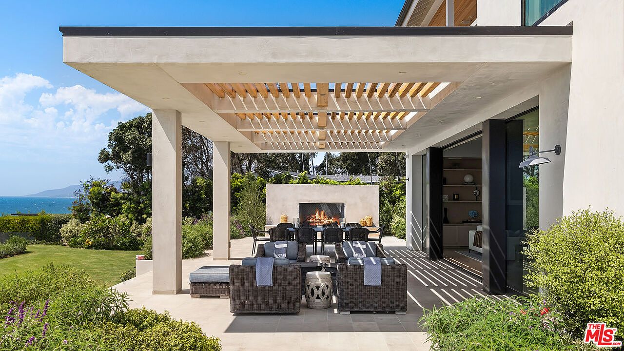 This luxurious outdoor living space features a covered patio with a modern pergola, a built-in fireplace, and comfortable lounge seating. The area seamlessly transitions from the indoor living space through large sliding glass doors, offering a stunning view of the ocean and lush landscaping. The design emphasizes a sophisticated, open-air lifestyle perfect for entertaining in a coastal setting.