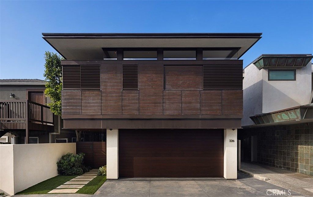 This modern, two-story residence features a striking facade with horizontal dark wood siding and a prominent, flat-roofed architectural design. The ground level is dominated by a wide, dark-toned garage door, while a small walkway with stone pavers leads to a side entrance. The clean lines and minimalist aesthetic create a sophisticated, contemporary curb appeal.