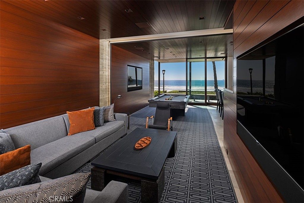 This sophisticated living area features rich wood-paneled walls and a geometric patterned rug that leads the eye toward a stunning ocean view. The space is furnished with a plush gray sofa, a dark coffee table, and a pool table positioned near large glass doors that open to the outdoors. The combination of warm wood tones and the expansive coastal backdrop creates a luxurious and serene atmosphere.