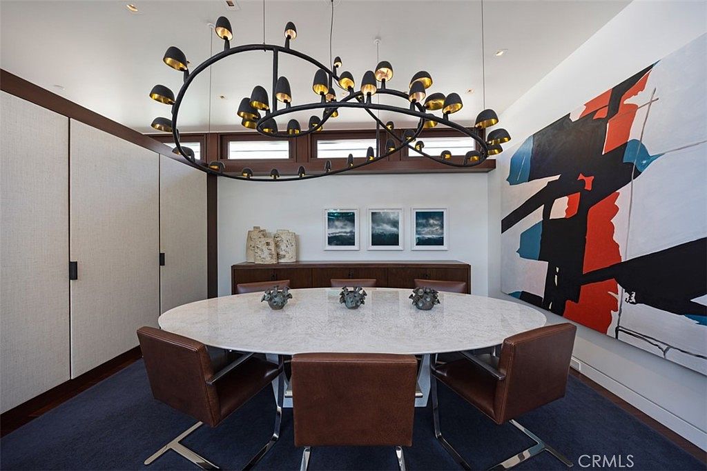 This sophisticated dining room features a large, oval-shaped stone-topped table surrounded by brown leather chairs, set against a deep blue area rug. A dramatic, multi-light sculptural chandelier serves as the focal point above the table, while a large, abstract mural with bold red and blue accents adorns the wall. The space is completed by a dark wood sideboard and a series of three framed landscape photographs, creating a refined and artistic atmosphere.