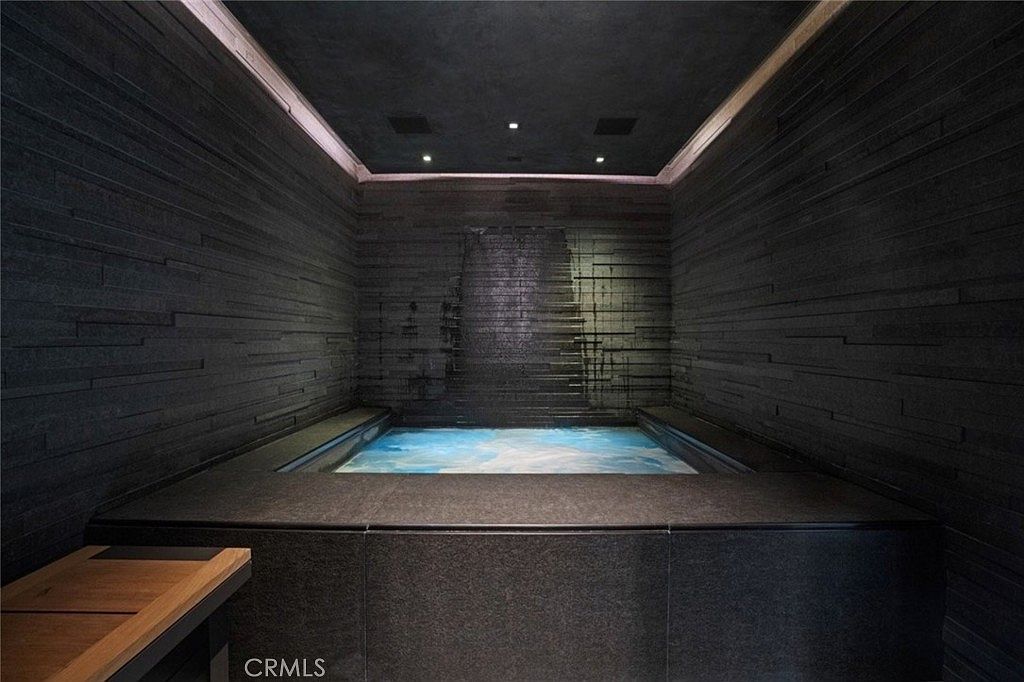 This luxurious indoor spa area features a dark, textured stone-clad wall with a built-in water feature cascading into a glowing, blue-lit pool. The room is framed by dark, horizontal stone paneling and recessed ceiling lighting, creating a moody, high-end atmosphere. A wooden bench sits in the foreground, providing a warm contrast to the sleek, dark aesthetic of the space.