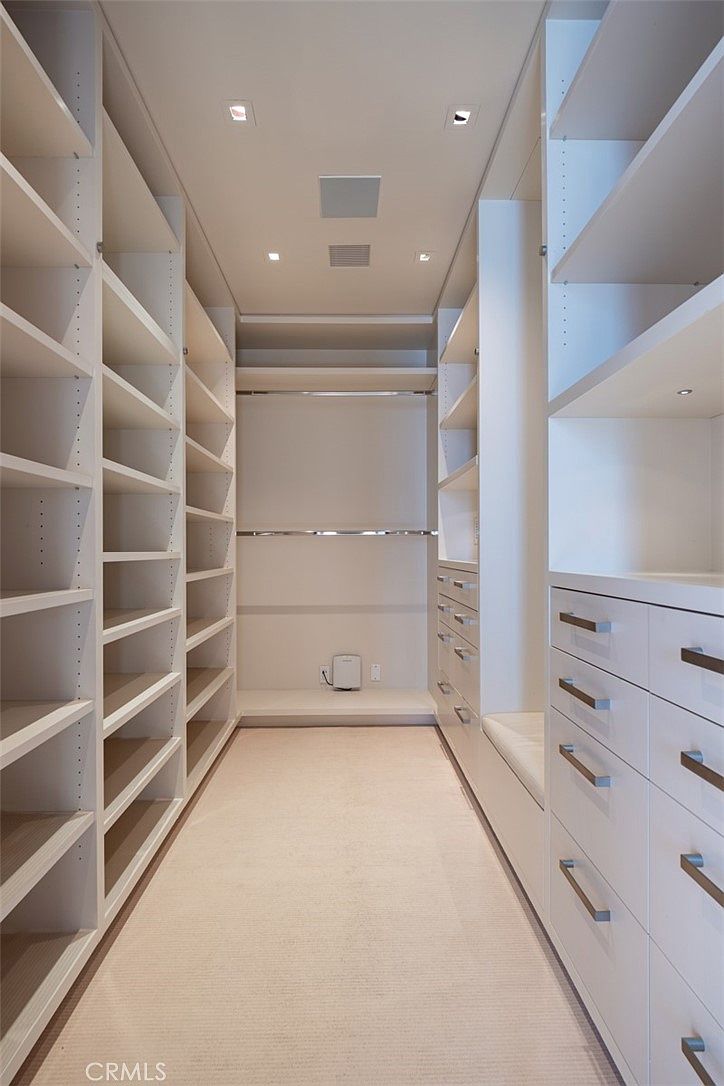 This spacious, symmetrical walk-in closet features extensive white cabinetry with open shelving and a bank of drawers on the right side. The design includes two levels of hanging rods at the back, a built-in bench, and recessed ceiling lighting that creates a bright, organized atmosphere. The neutral carpet and clean lines provide a minimalist and highly functional aesthetic.