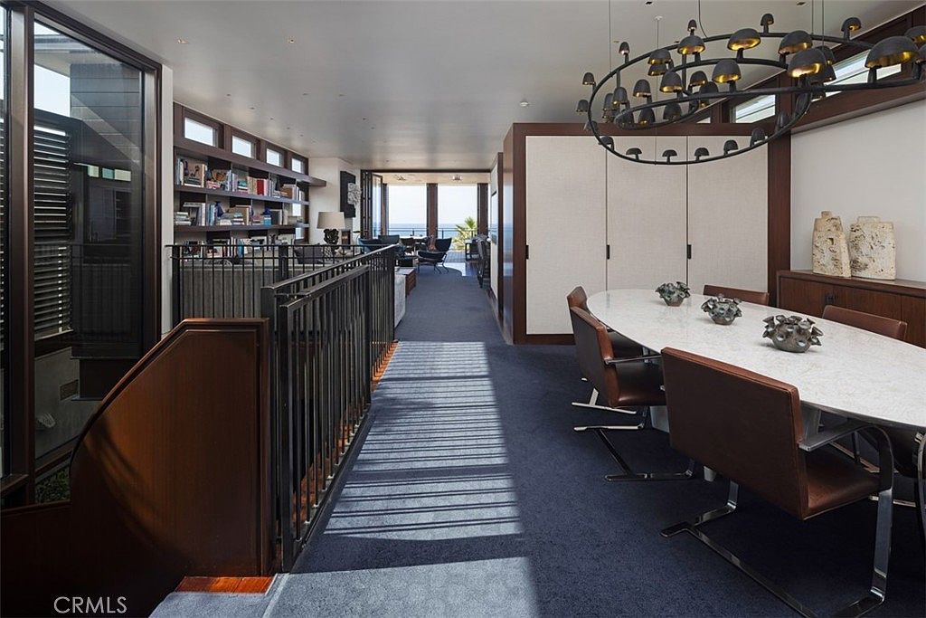 This sophisticated dining area features a long, oval-shaped table with a light-colored stone top, surrounded by brown leather chairs. A unique, multi-bulb circular chandelier hangs above the table, while a dark metal railing separates the dining space from a sunken living area with built-in bookshelves. The open-concept layout leads the eye toward large windows at the far end, providing a clear view of the ocean beyond.