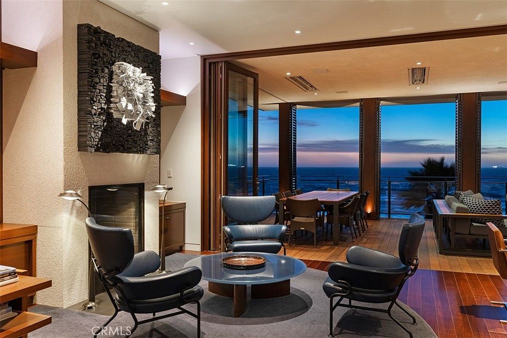 This sophisticated living area features a central seating arrangement with dark leather lounge chairs surrounding a circular glass-topped coffee table. A prominent textured art piece hangs above the fireplace, which is set into a neutral-toned wall. Large glass doors open onto an expansive outdoor deck, providing an unobstructed view of the ocean at twilight.