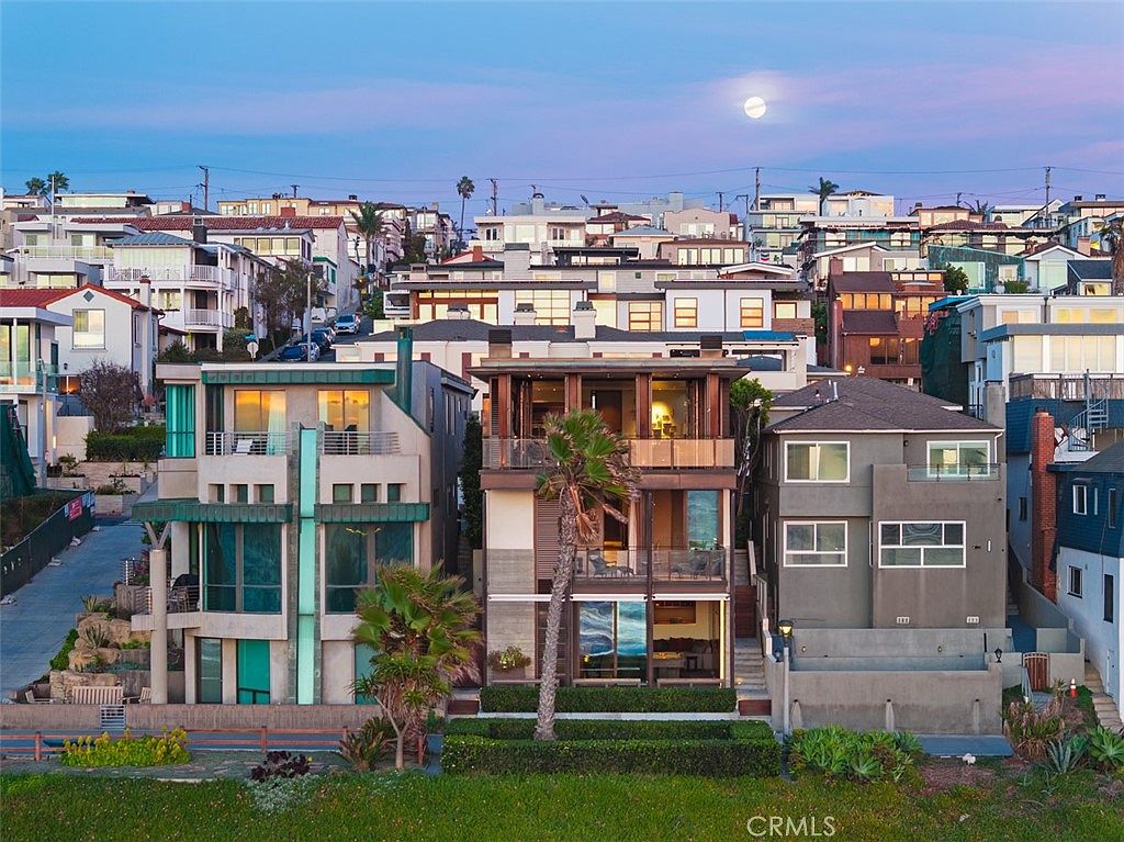This eye-level exterior shot captures a row of multi-story coastal homes at dusk, with a prominent moon visible in the sky. The central residence features a modern design with stacked balconies, large glass windows, and a palm tree framing the facade. The surrounding neighborhood is densely packed with various architectural styles, creating a vibrant and upscale beachside atmosphere.