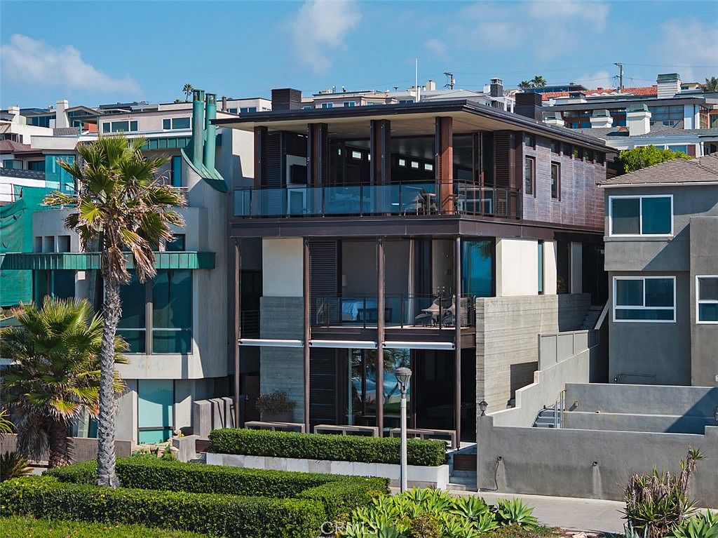 This modern, multi-level coastal residence features a striking architectural design with clean lines and a prominent use of glass and dark structural framing. The home includes expansive balconies on the upper levels that provide seamless indoor-outdoor living, overlooking a manicured hedge and a paved walkway. The structure is nestled among neighboring homes, showcasing a contemporary aesthetic that emphasizes openness and natural light.