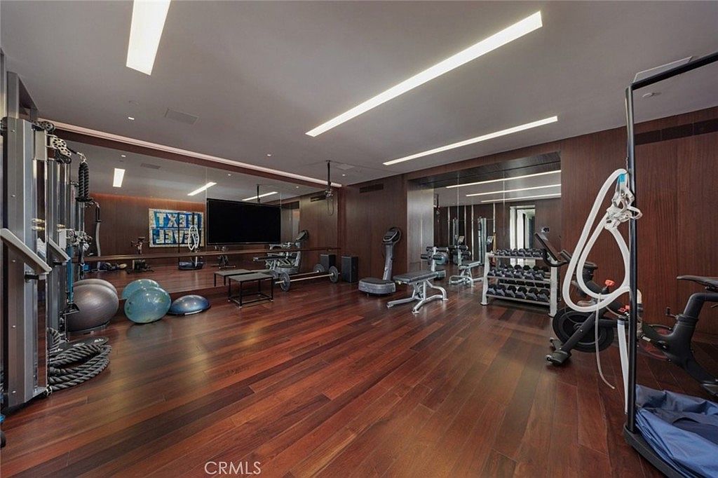 This spacious home gym features rich, dark wood flooring and expansive wall-to-wall mirrors that enhance the sense of depth in the room. The space is well-equipped with a variety of fitness gear, including exercise balls, a weight rack, a television, and a stationary bike. Recessed linear lighting integrated into the ceiling provides a clean, modern aesthetic, making the room feel professional and inviting.