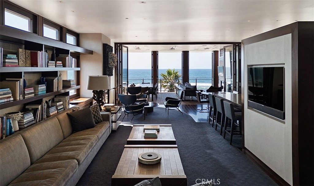 This expansive, open-concept living area features a sophisticated, modern aesthetic with a long, plush sofa and dual wooden coffee tables centered on a dark area rug. A wall of built-in shelving provides ample storage and display space, while the far end of the room opens completely to a deck with panoramic ocean views through large glass doors. The space is designed for seamless indoor-outdoor living, characterized by clean lines, dark wood tones, and a serene, coastal-inspired atmosphere.