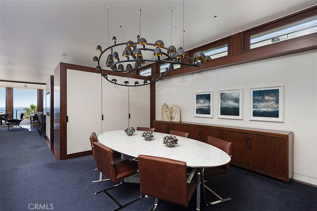 This sophisticated dining room features an oval table with a light-colored stone top, surrounded by six brown leather chairs. A large, circular, multi-light chandelier serves as a dramatic focal point above the table, while a long wooden sideboard sits against the wall beneath three framed landscape prints. The room is finished with dark carpeting and a wall of tall, horizontal windows that provide natural light and a glimpse of the ocean view beyond.
