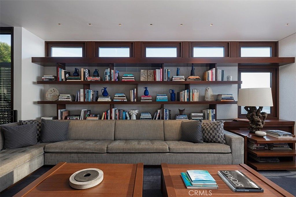 This sophisticated living room features a large, neutral-toned sectional sofa positioned in front of an expansive wall of built-in wooden shelving. The shelving unit incorporates a series of clerestory windows that allow natural light to filter into the space, creating an airy and architectural aesthetic. Two wooden coffee tables sit in the foreground, while a unique, textured table lamp adds a sculptural element to the side table.