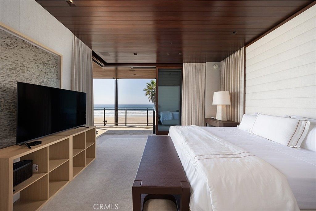 This primary bedroom features a minimalist, modern aesthetic with a large bed facing a wall-mounted television and a light wood media console. The room is defined by its warm wood-paneled ceiling and expansive glass doors that open directly onto a private balcony overlooking the ocean. The neutral color palette and clean lines create a serene, high-end atmosphere that emphasizes the seamless connection between the indoor living space and the coastal environment.