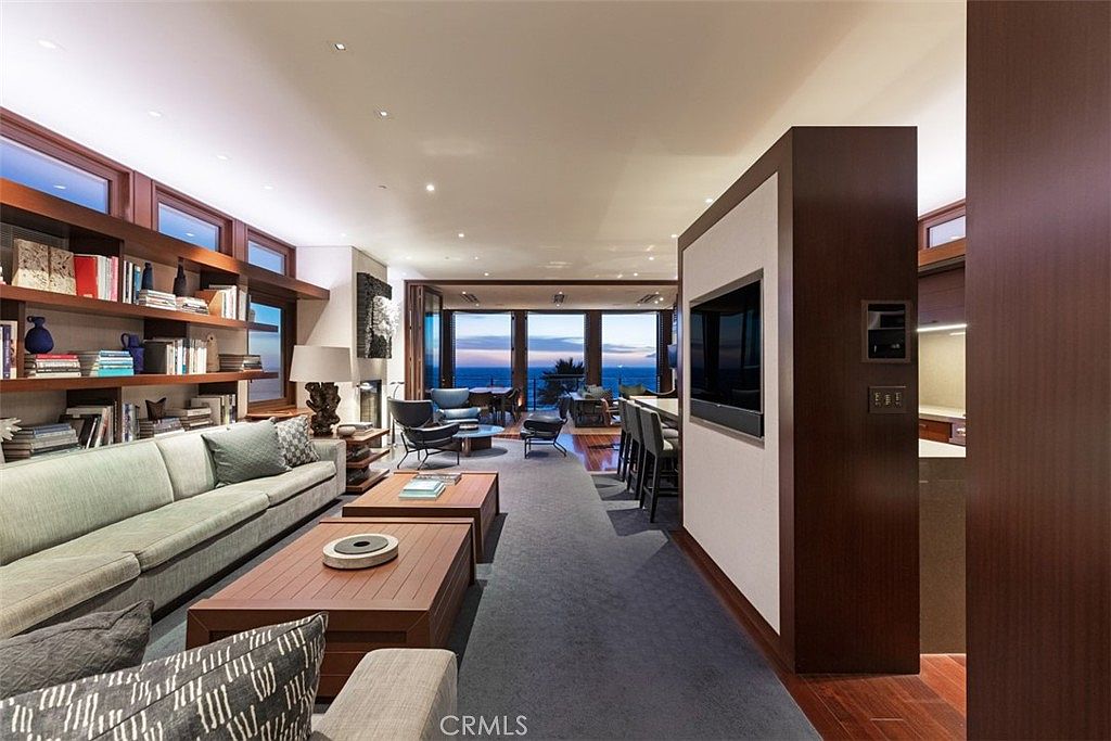 This sophisticated open-concept living area features a long, neutral-toned sofa facing a pair of low-profile wooden coffee tables. A prominent dark wood partition wall houses a television, separating the living space from the adjacent kitchen area, while a wall of built-in shelving provides ample display space. The room flows toward a set of large glass doors that reveal a stunning ocean view at dusk, creating a seamless transition between the refined interior and the coastal horizon.